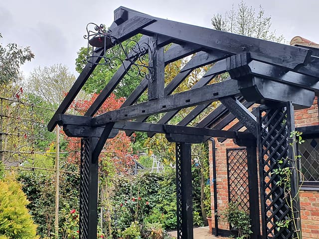 Close-up of a black painted timber pergola with a decorative wrought-iron bracket and autumn foliage in a Surrey garden